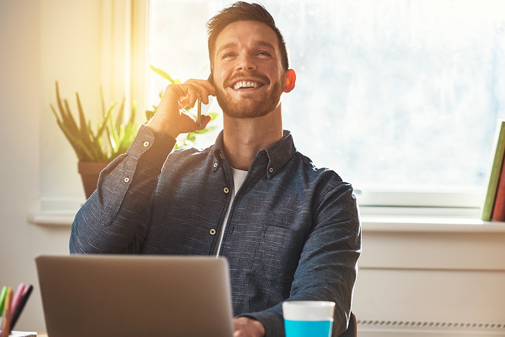 Man zit achter laptop en belt met zijn mobiele telefoon.